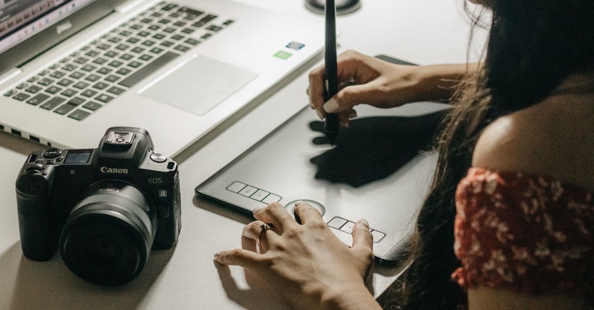 A designer drawing on a pen tablet with a stylus, beside a laptop and a camera on a white desk.
